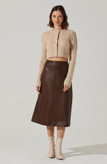 A woman is wearing a beige, long-sleeved, cable-knit cardigan with a button-down front. The cardigan is cropped, ending above the waist. She pairs it with a high-waisted, dark brown leather midi skirt that reaches mid-calf. She completes the outfit with beige ankle boots.