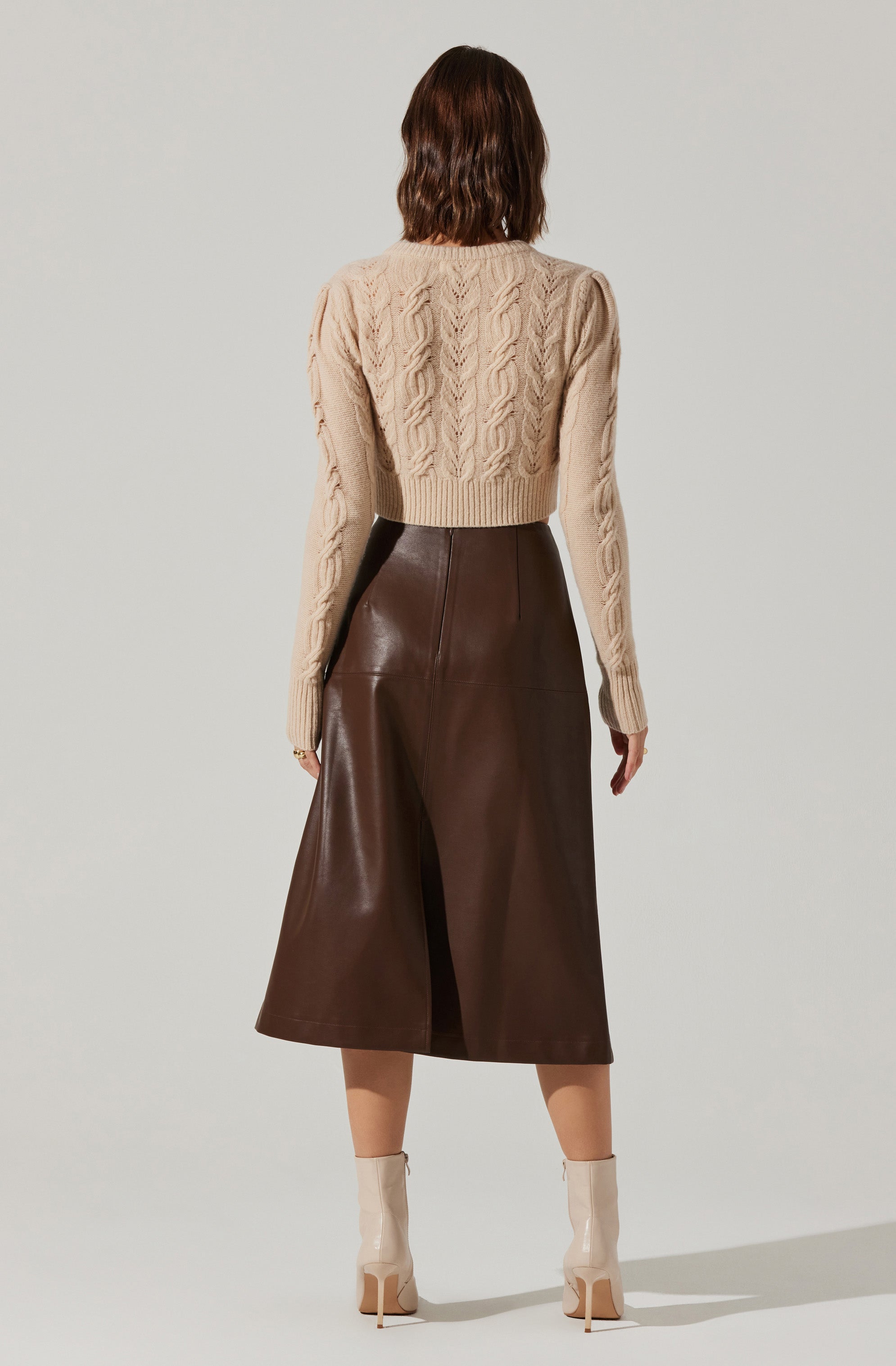 A person is wearing a beige cable-knit sweater with a high neckline and long sleeves. They are also wearing high-waisted, wide-leg brown leather culottes that fall below the knees. The outfit is complemented by light-colored ankle boots with high heels.