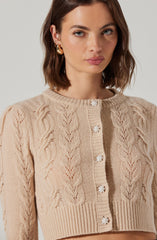 A woman is wearing a beige cropped cardigan with a cable knit pattern. The cardigan features a round neckline and decorative floral buttons down the front. She has shoulder-length brown hair and is accessorized with gold hoop earrings.