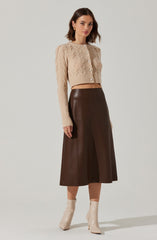 A woman is wearing a beige, cropped, cable-knit cardigan with long sleeves and decorative buttons down the front. She pairs it with a high-waisted, dark brown, midi-length skirt and light-colored ankle boots with a pointed toe and a stiletto heel.