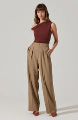 A woman is wearing a sleeveless, asymmetrical burgundy top with a draped design at the neckline. She pairs it with high-waisted, wide-leg beige trousers that have pleats at the front and side pockets. The outfit is completed with pointed-toe shoes.