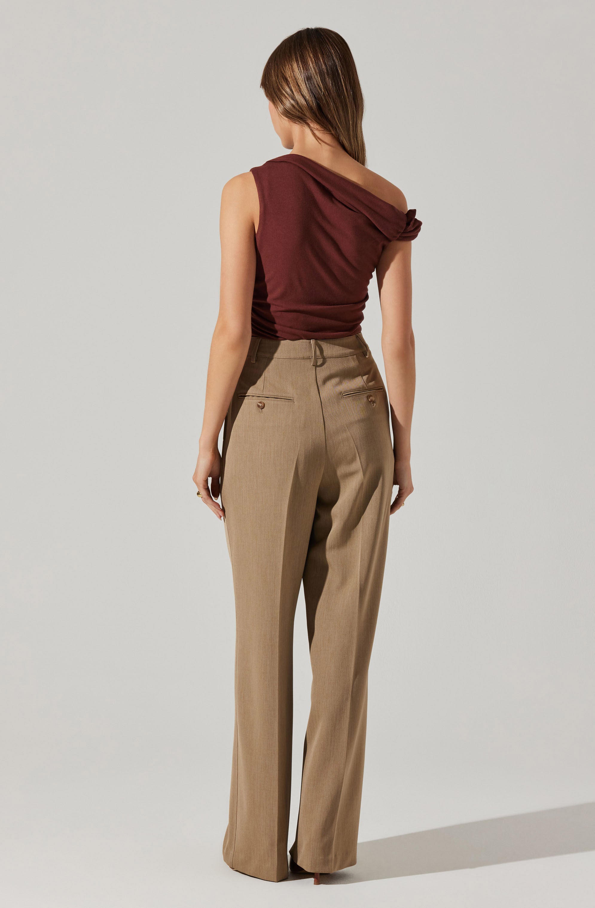 The image shows a woman wearing a sleeveless, dark red, one-shoulder top with a gathered detail on the shoulder. She is also dressed in high-waisted, beige, wide-leg trousers with two back pockets featuring button closures.