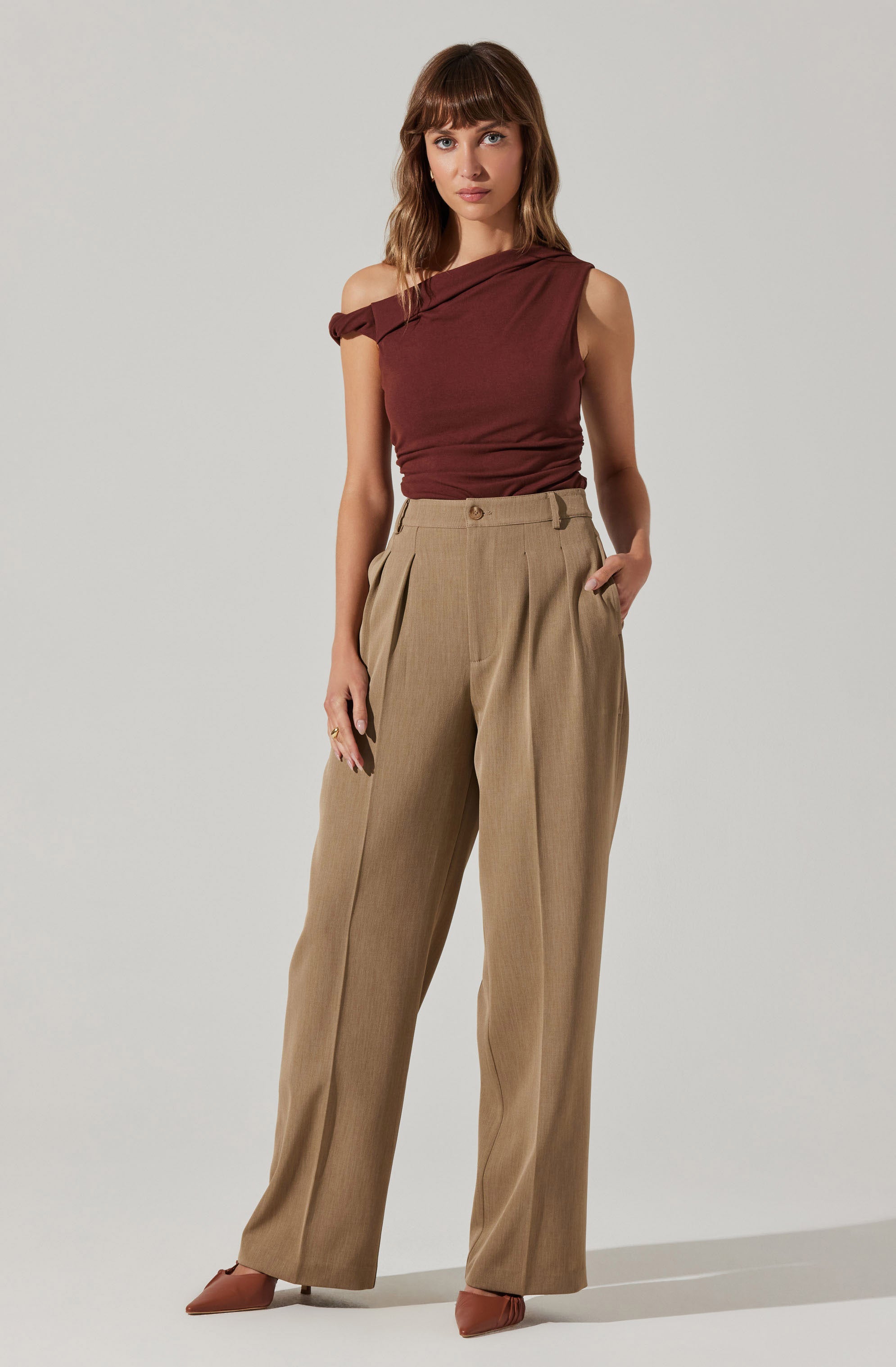 A woman is wearing a sleeveless, off-the-shoulder top in a maroon color with a slight wrap or drape detail at one shoulder. She is also dressed in high-waisted, wide-leg beige trousers with pleats at the front and side pockets. She completes her look with pointed toe shoes in a matching brown shade.