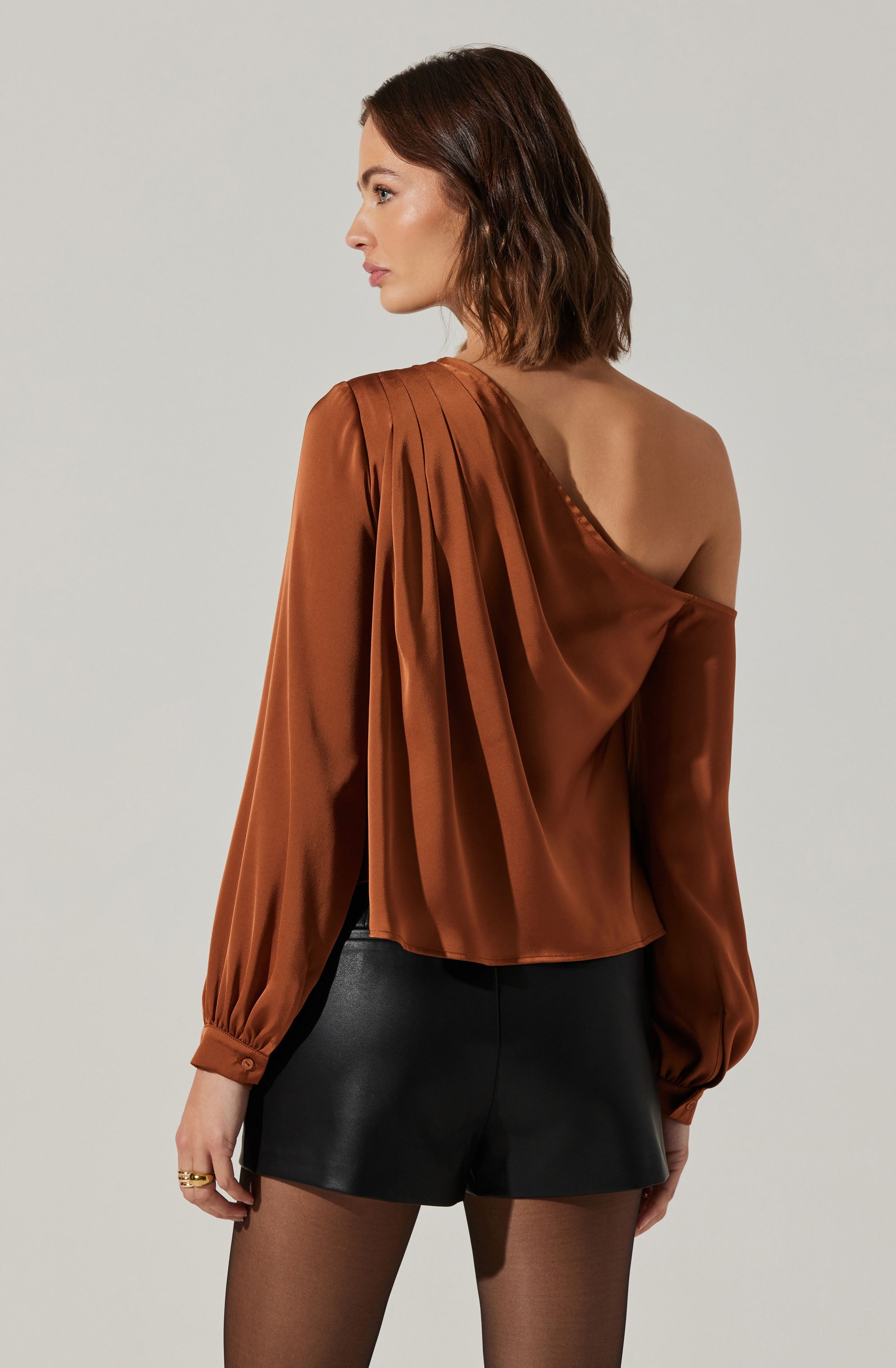 A woman is wearing a shiny, satin-style, rust-colored, one-shoulder blouse with pleats on the shoulder and long, puffed sleeves gathered at the wrists. She pairs it with black leather shorts and sheer black tights.