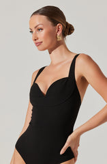 A woman is wearing a black sleeveless bodysuit with wide straps and a sweetheart neckline. Her hair is styled in a neat bun, and she is accessorized with gold rectangular earrings.