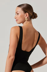 A woman with her hair styled in a low bun is wearing a black sleeveless leotard with wide straps and a deep scoop back. She has on bold, gold statement earrings.