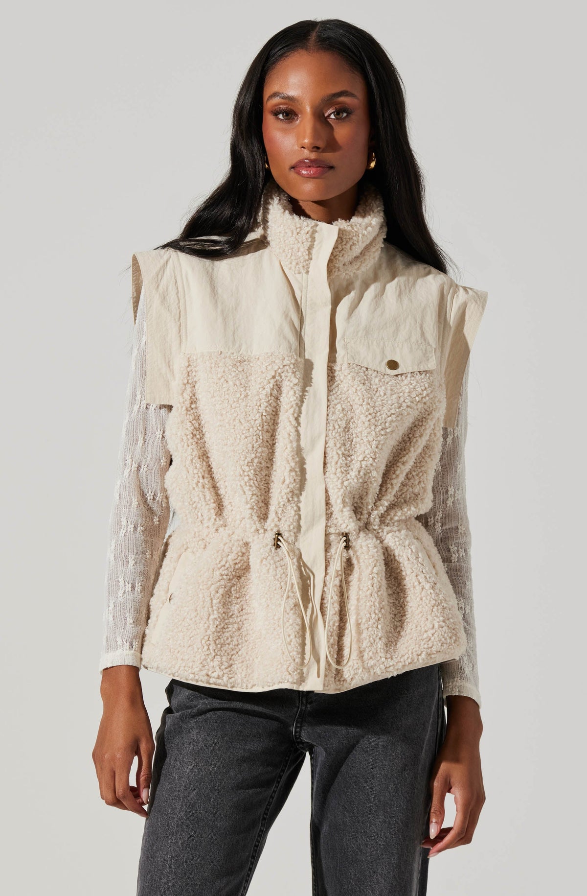 A woman is wearing a sleeveless, cream-colored fleece vest with a high collar and a front zipper. The vest features a drawstring waist, giving it a slightly cinched appearance, and has a small pocket with a button flap on the left chest. She is also wearing a long-sleeved, white lace top underneath the vest. She pairs this with dark gray jeans.