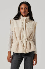 A woman is wearing a sleeveless, cream-colored fleece vest with a high collar and a front zipper. The vest features a drawstring waist, giving it a slightly cinched appearance, and has a small pocket with a button flap on the left chest. She is also wearing a long-sleeved, white lace top underneath the vest. She pairs this with dark gray jeans.