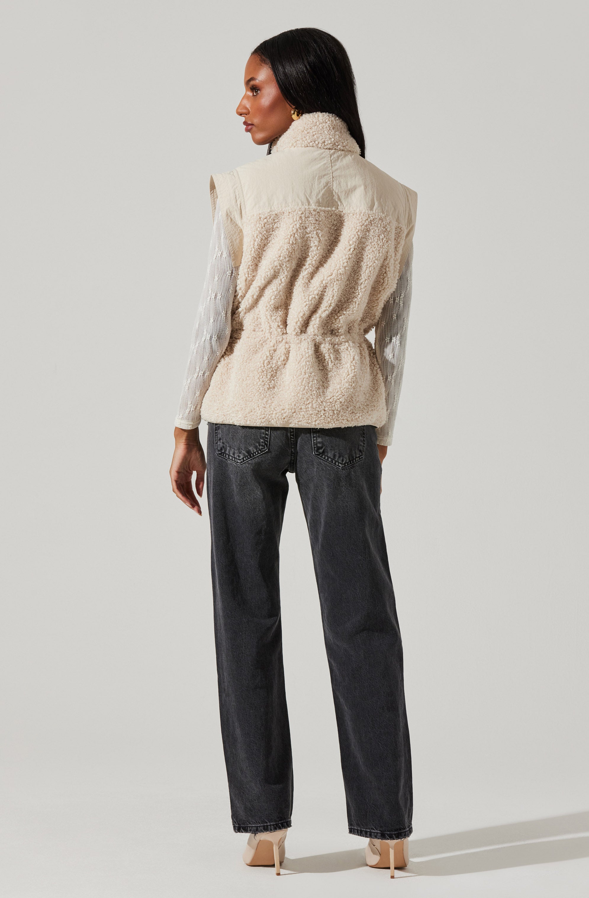 A woman is wearing a sleeveless, cream-colored vest with a fluffy, textured fabric. Underneath, she has on a long-sleeved, white blouse with subtle vertical stripes. She pairs these with high-waisted, dark gray jeans and beige high-heeled shoes.