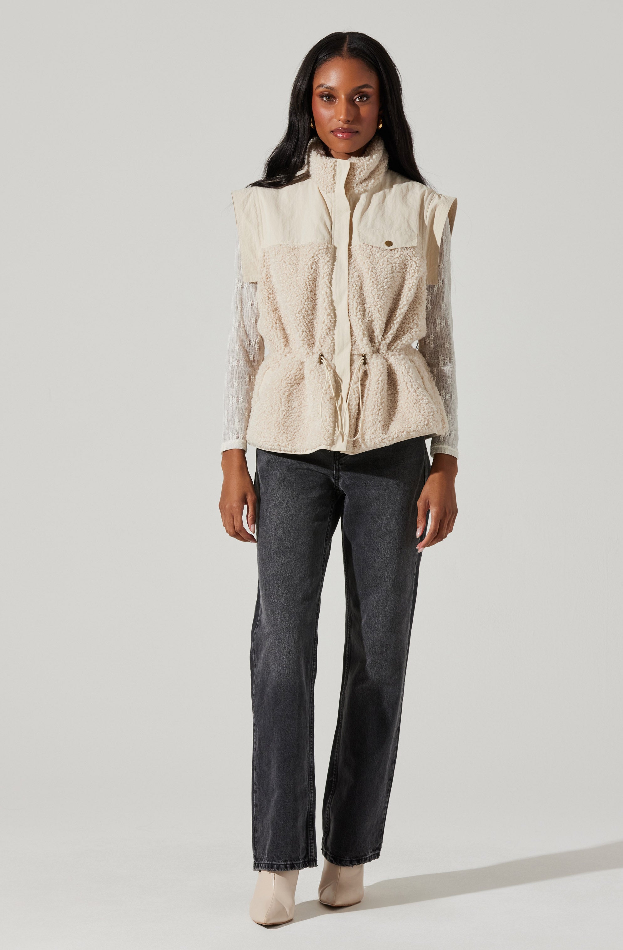 A woman is wearing a sleeveless, cream-colored vest with textured paneling and a high collar. Underneath, she has on a long-sleeve, white shirt made of a sheer fabric that extends past the vest’s shoulders. She pairs this with high-waisted, straight-leg black jeans and light-colored ankle boots.