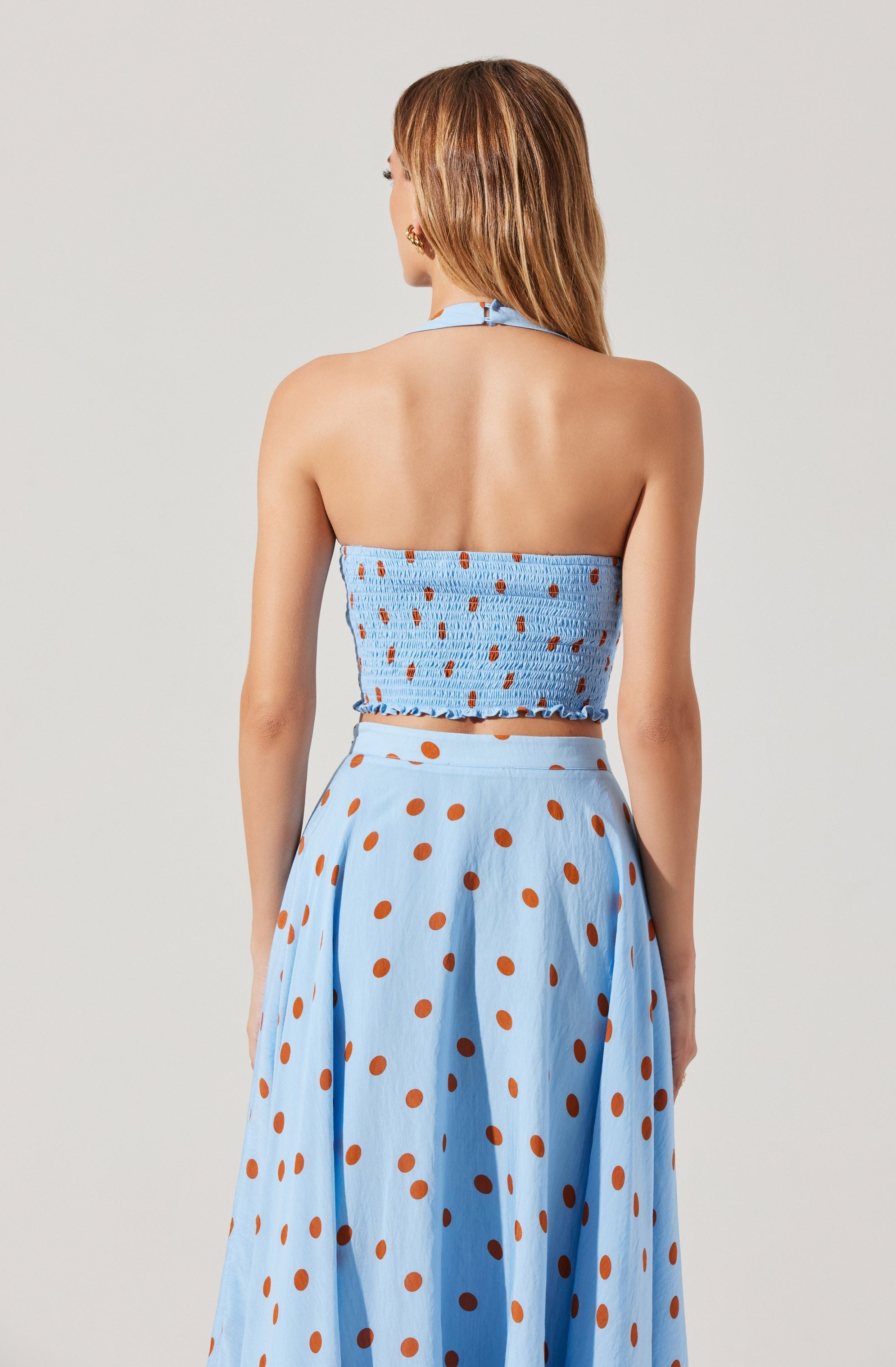 A woman is wearing a matching light blue two-piece outfit with brown polka dots. The top is a strapless, smocked bandeau style with a ruffled hem and a halter strap tied at the neck. The high-waisted skirt is long and flowing, featuring the same brown polka dots throughout.