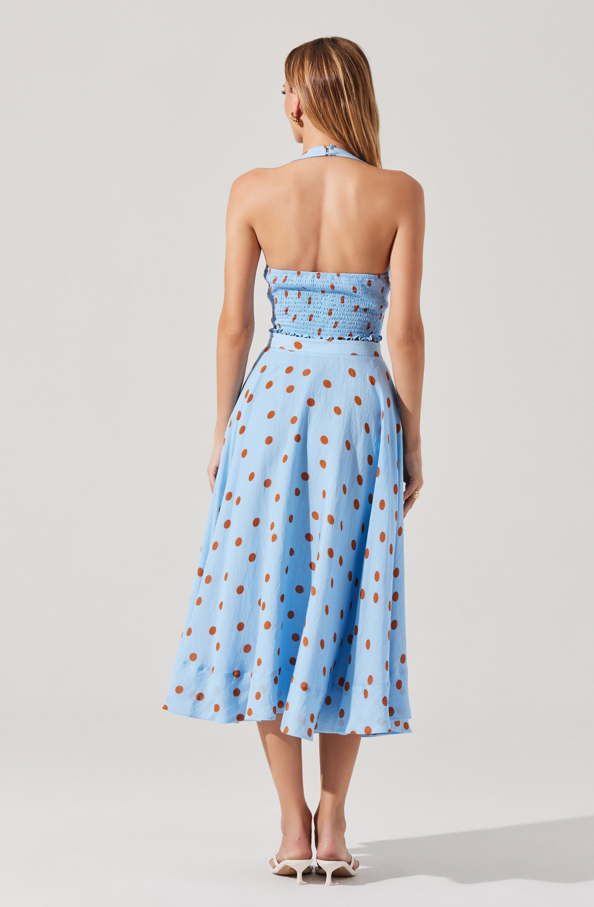 A woman is wearing a light blue halter top with elastic shirring at the back and a matching high-waisted, full-length skirt. The skirt features a brown polka dot pattern throughout. She is also wearing white high-heeled shoes.