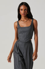 A woman is wearing a dark gray sleeveless jumpsuit with thin, vertical pinstripes. The top has a square neckline and is fitted at the waist. The pants are high-waisted with a pleated front and a tailored, wide-leg cut. She accessorizes with gold hoop earrings, a chunky gold chain necklace, and rings on both hands. Her long, dark hair is styled straight and loose.