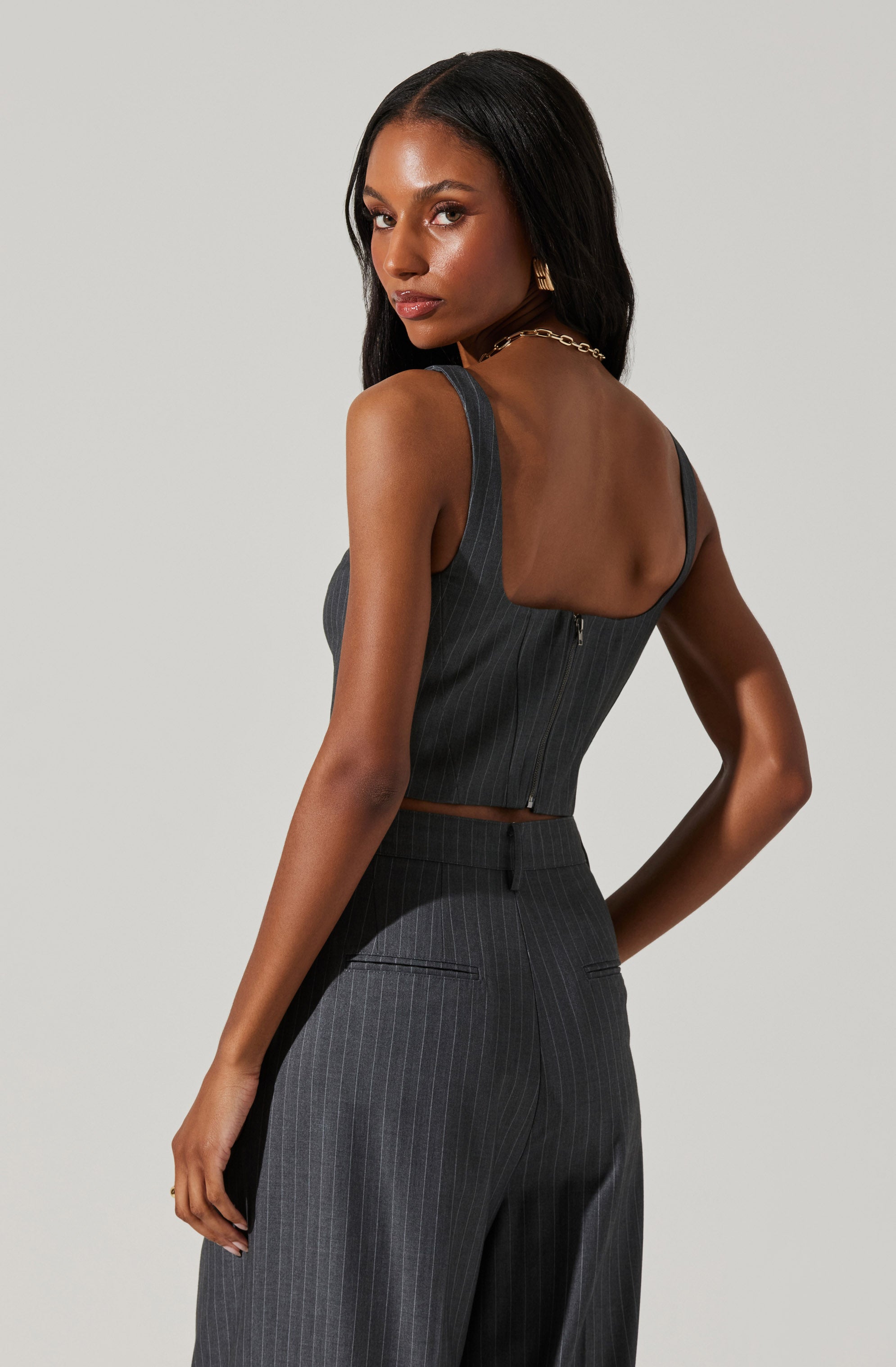 A woman is wearing a dark gray pinstripe two-piece outfit. The top is a sleeveless, fitted crop top with wide straps and a back zipper closure. The high-waisted pants are tailored with a straight-leg fit, featuring belt loops and two small horizontal slit pockets on the back.