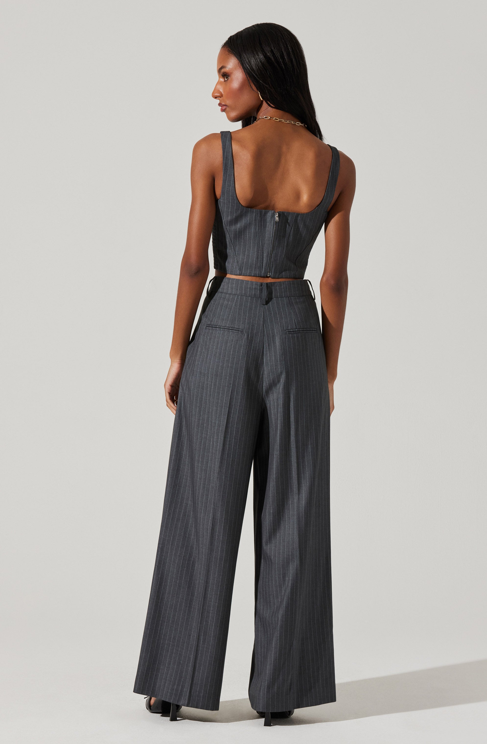 A woman is wearing a matching dark gray pinstripe sleeveless top with wide straps and high-waisted, wide-leg trousers. The top has a zipper down the back, and the trousers feature belt loops and two back pockets.