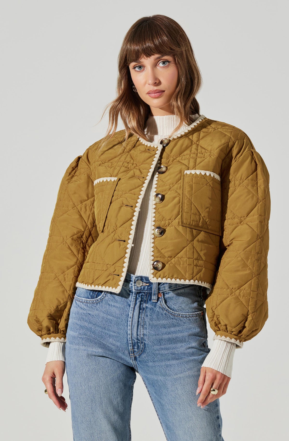 A woman is wearing a mustard yellow, quilted, cropped jacket with large buttons down the front and decorative white scalloped trim along the collar, pockets, and hem. Underneath, she has on a white ribbed sweater with ribbing at the cuffs. She pairs this with high-waisted, light-wash jeans.