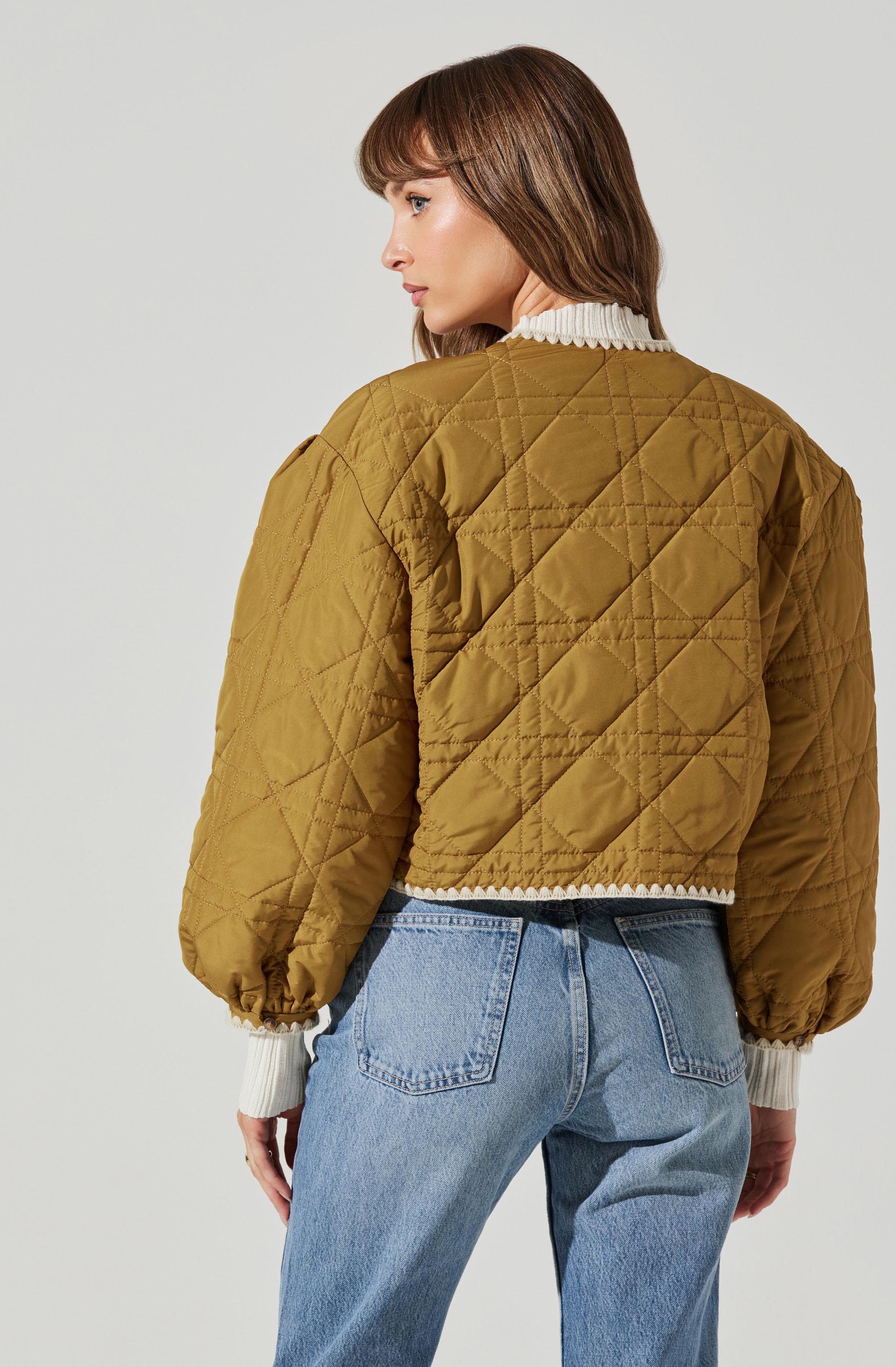 A woman is wearing a quilted, olive-green jacket with a slightly cropped fit. The jacket features puffy, long sleeves with elasticized cuffs, and a diamond-shaped quilted pattern throughout. She is also wearing a white, ribbed sweater underneath, with the collar and cuffs visible.