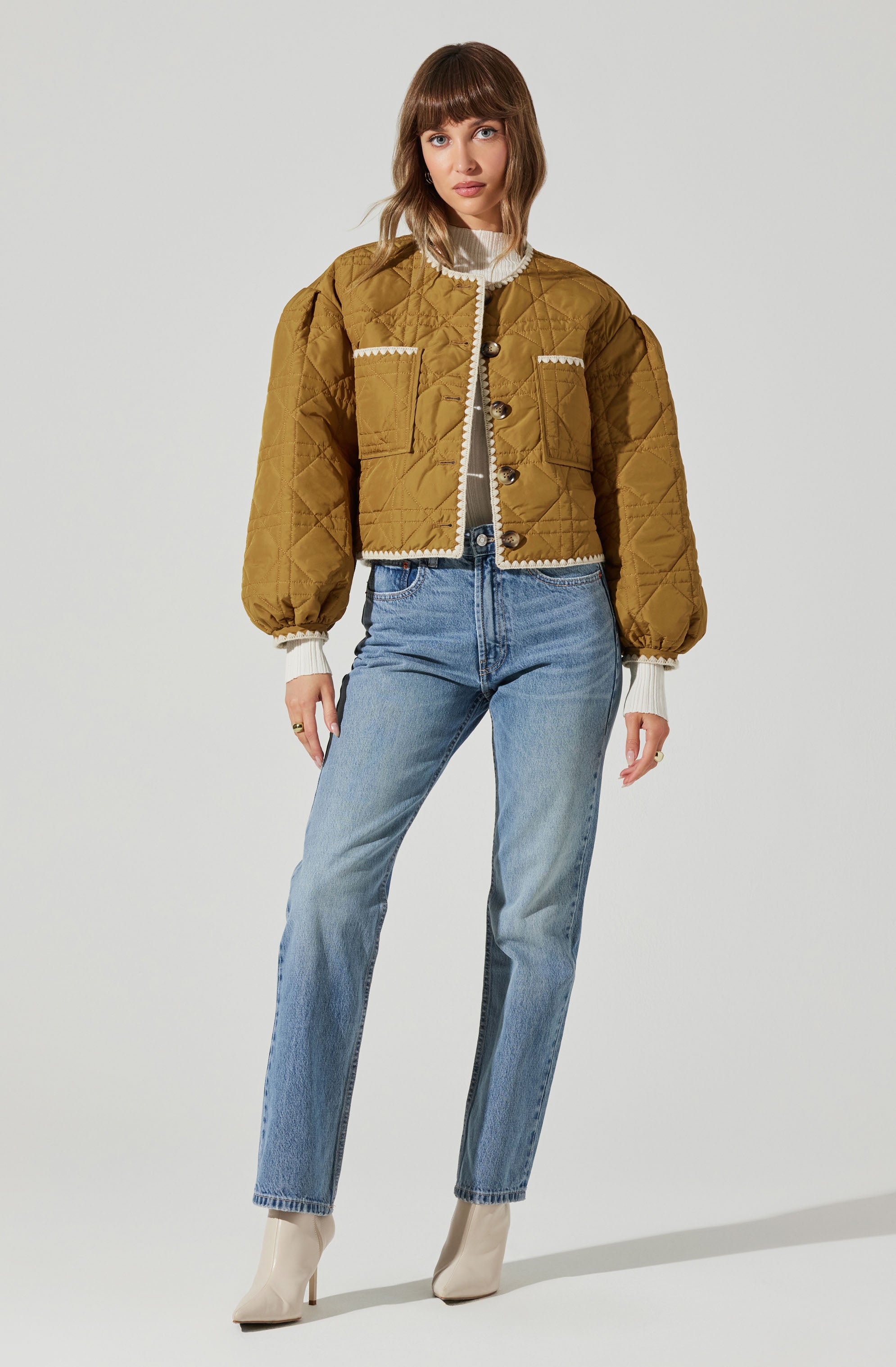 A woman is wearing a mustard-colored, quilted, cropped jacket with puffed sleeves, button closure, and decorative white piping details on the pockets and edges. She has on a white turtleneck sweater underneath, paired with high-waisted light blue jeans and beige ankle boots.