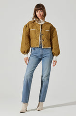 A woman is wearing a mustard-colored, quilted, cropped jacket with puffed sleeves, button closure, and decorative white piping details on the pockets and edges. She has on a white turtleneck sweater underneath, paired with high-waisted light blue jeans and beige ankle boots.
