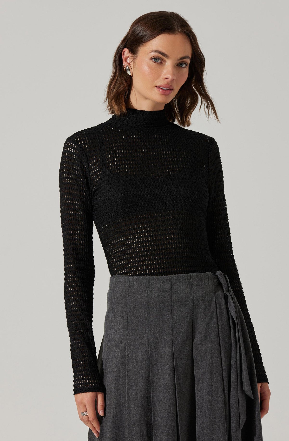 A woman is wearing a black, long-sleeved, high-neck sweater with a perforated, open-knit pattern. She pairs it with a high-waisted gray skirt that features vertical pleats and a matching fabric tie belt.