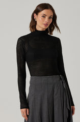 A woman is wearing a black, long-sleeved, high-neck sweater with a perforated, open-knit pattern. She pairs it with a high-waisted gray skirt that features vertical pleats and a matching fabric tie belt.