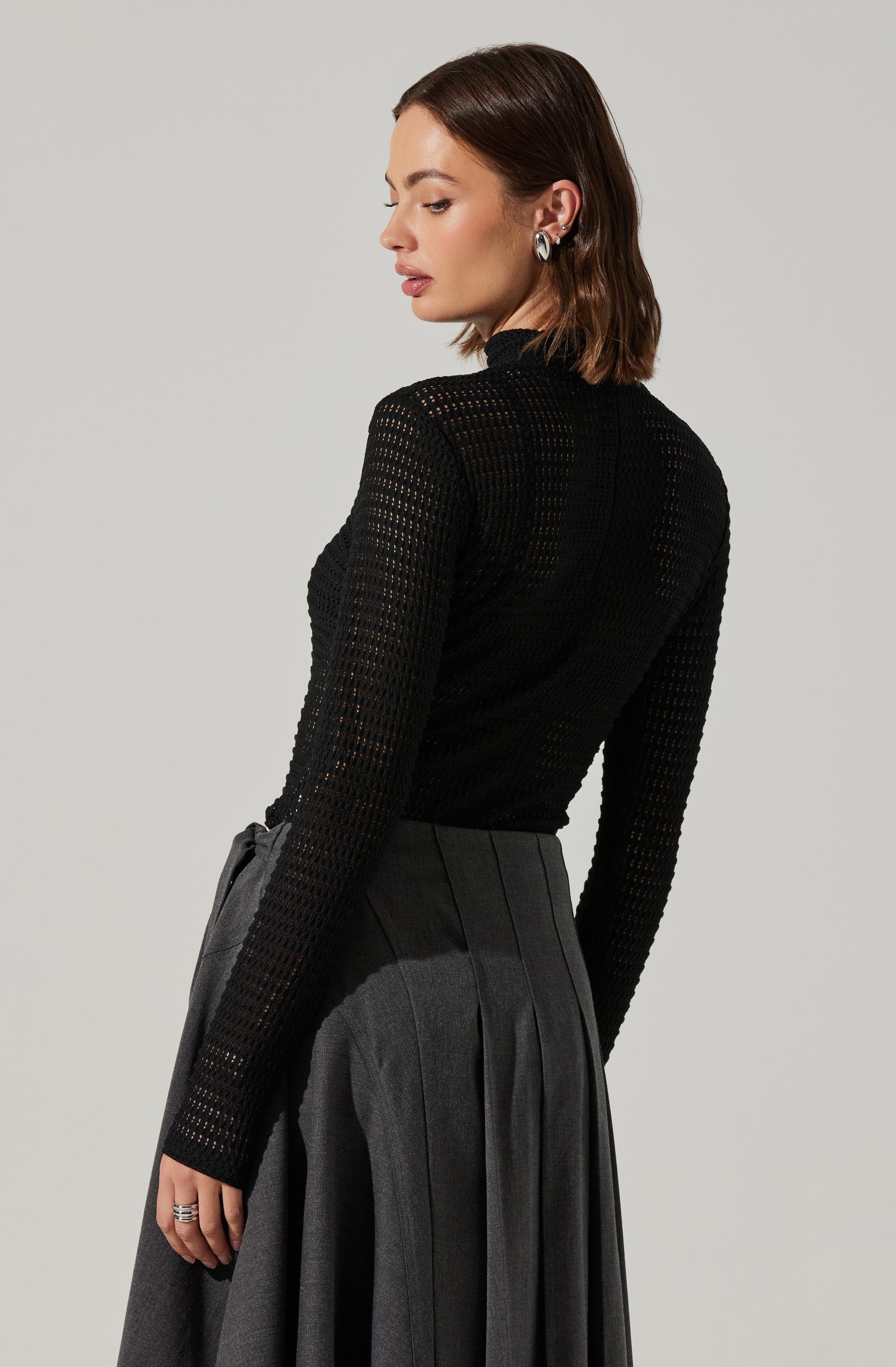 A woman is wearing a black, long-sleeved, open-knit top that appears slightly see-through, paired with a high-waisted, dark gray pleated skirt. She has short, wavy brown hair and is accessorized with silver hoop earrings.