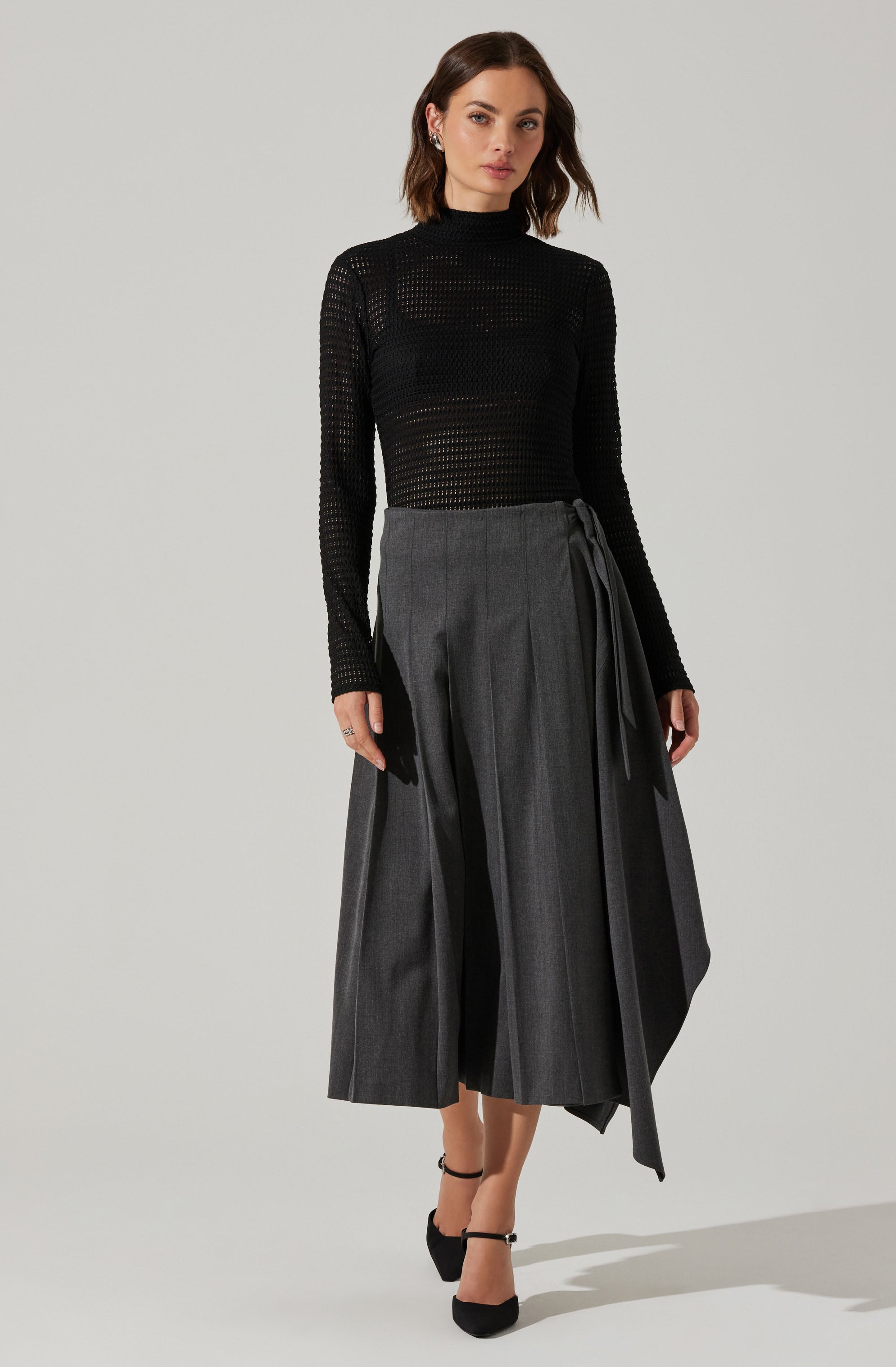 A woman is wearing a fitted black long-sleeve top with a high neckline, made of a see-through, open-knit fabric. She pairs it with a high-waisted, wide-leg gray skirt that features vertical pleats and a tied sash detail on one side. She completes her outfit with black ankle-strap high heels.