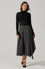 A woman is wearing a fitted black long-sleeve top with a high neckline, made of a see-through, open-knit fabric. She pairs it with a high-waisted, wide-leg gray skirt that features vertical pleats and a tied sash detail on one side. She completes her outfit with black ankle-strap high heels.