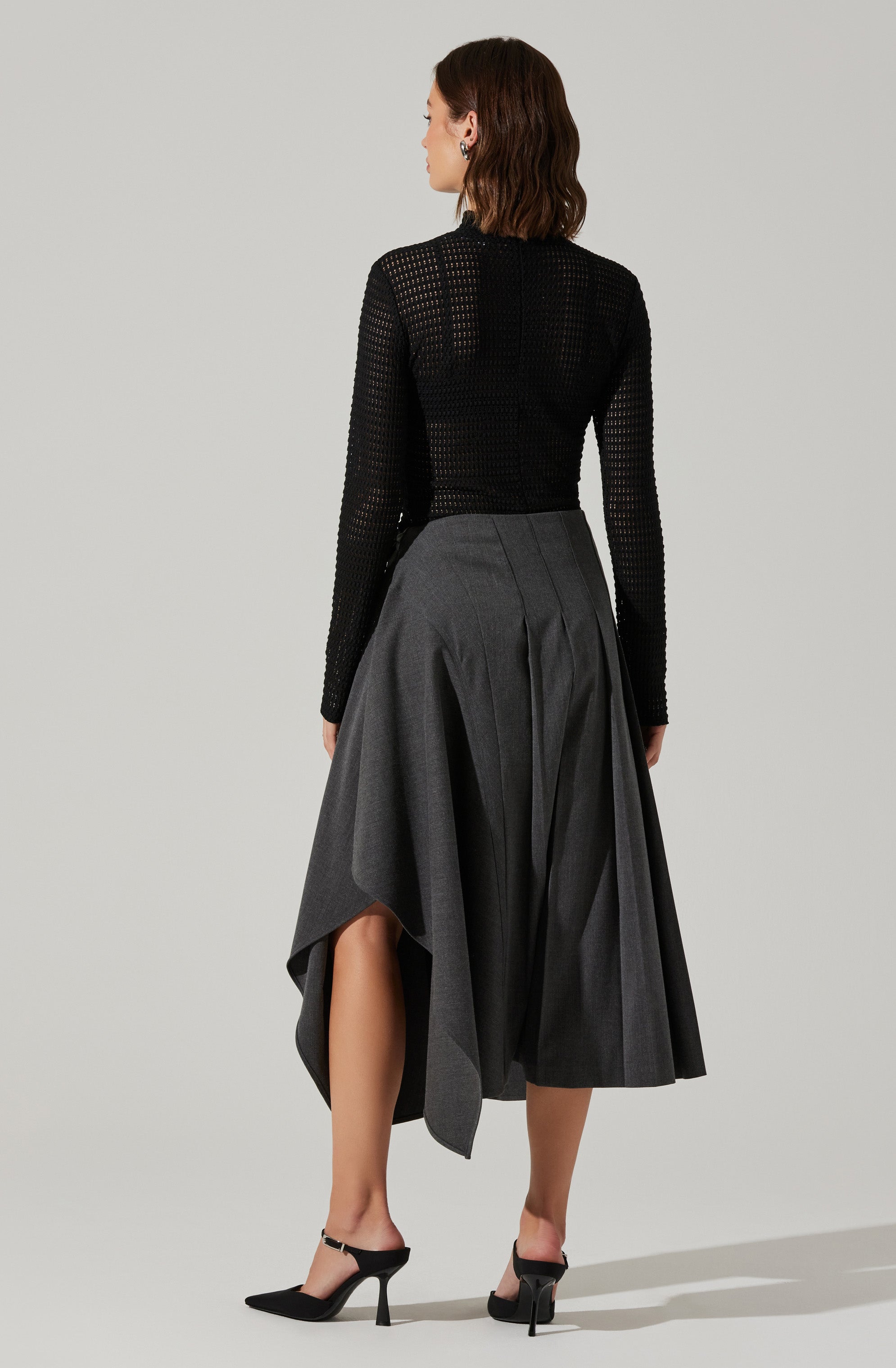 A woman is wearing a black, long-sleeved, open-knit sweater paired with a high-waisted, dark gray, asymmetrical pleated skirt that reaches below the knees on one side and above the ankle on the other. She completes the outfit with black high-heeled shoes featuring ankle straps.