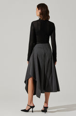 A woman is wearing a black, long-sleeved, open-knit sweater paired with a high-waisted, dark gray, asymmetrical pleated skirt that reaches below the knees on one side and above the ankle on the other. She completes the outfit with black high-heeled shoes featuring ankle straps.