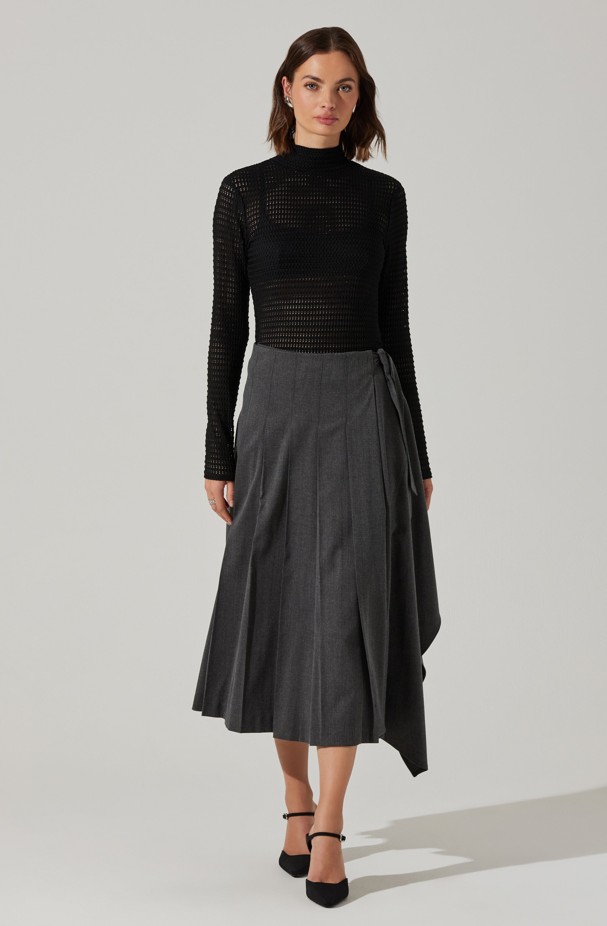 A woman is wearing a black, long-sleeved, semi-sheer knit top with a high neckline. She pairs it with a high-waisted, dark gray, pleated midi skirt that has a wrap style with a tie on the side. She completes her outfit with black ankle-strap high heels.