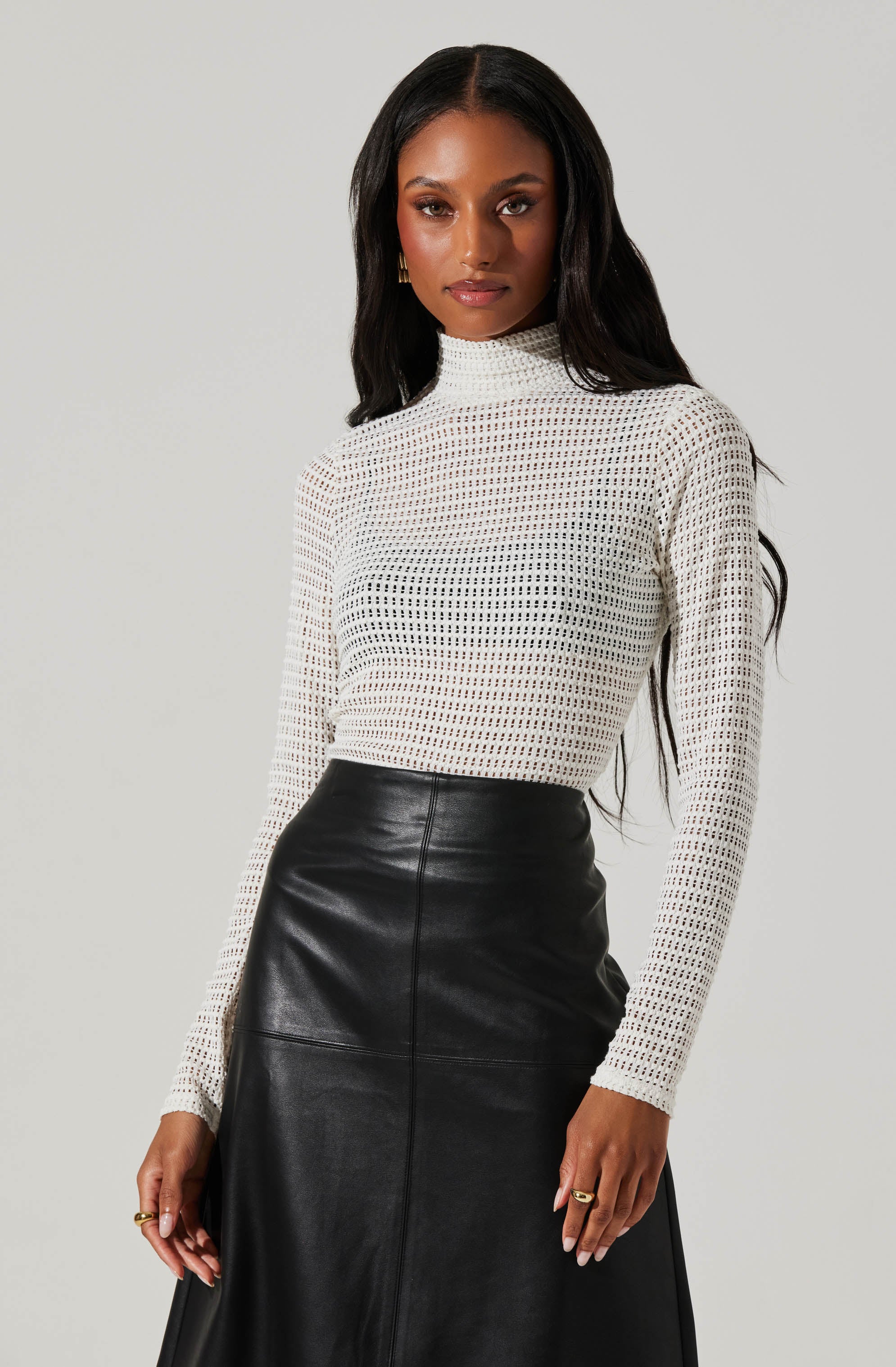 A woman is wearing a white, long-sleeved, high-neck knit sweater with a grid-like open weave pattern. She pairs it with a high-waisted black leather skirt that extends below her knees. She has long, dark hair and is accessorized with small gold earrings and rings.