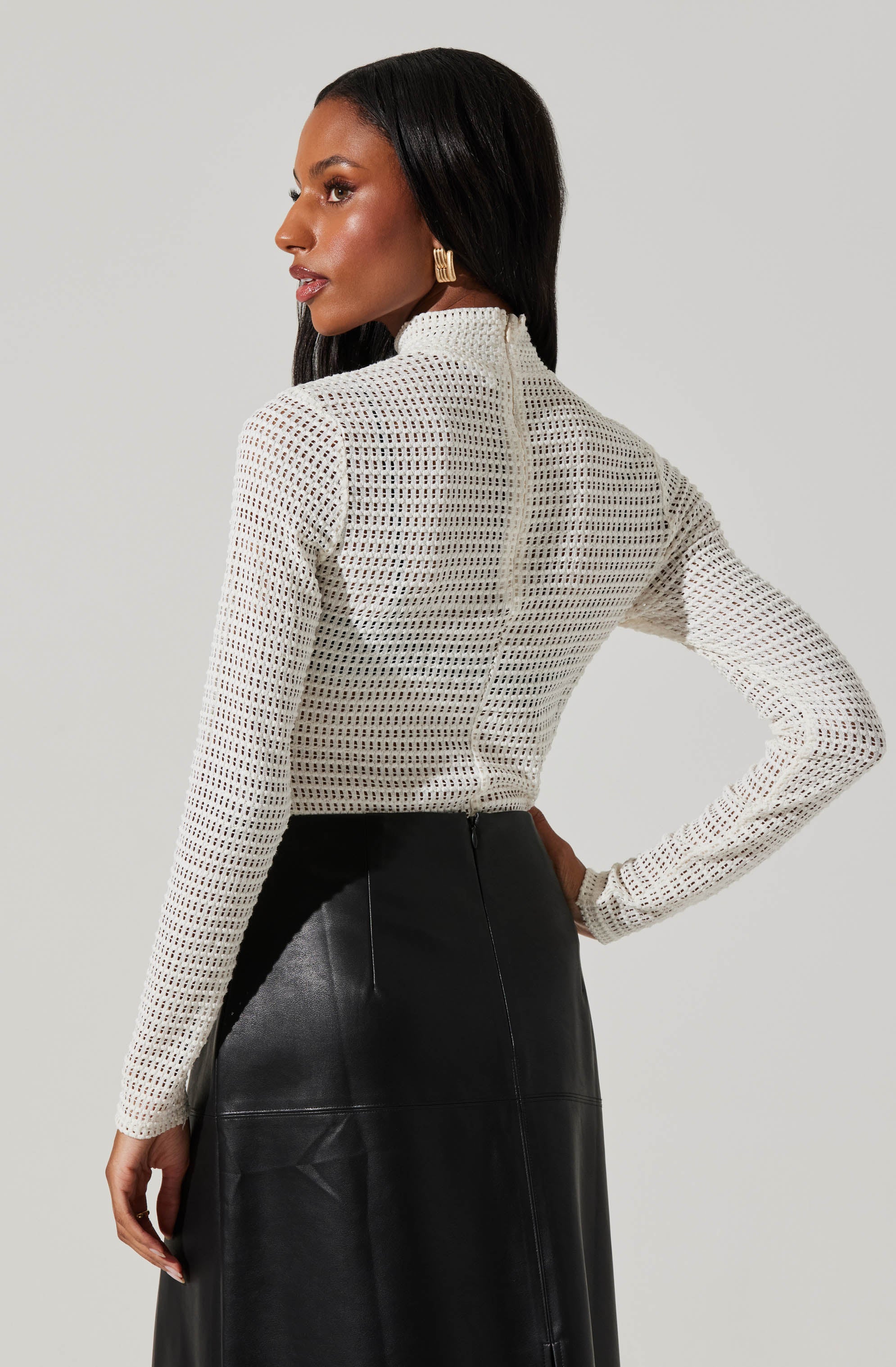 A woman is wearing a white, long-sleeved, high-neck crochet top with a see-through, grid-like pattern. She pairs it with a high-waisted black leather skirt.