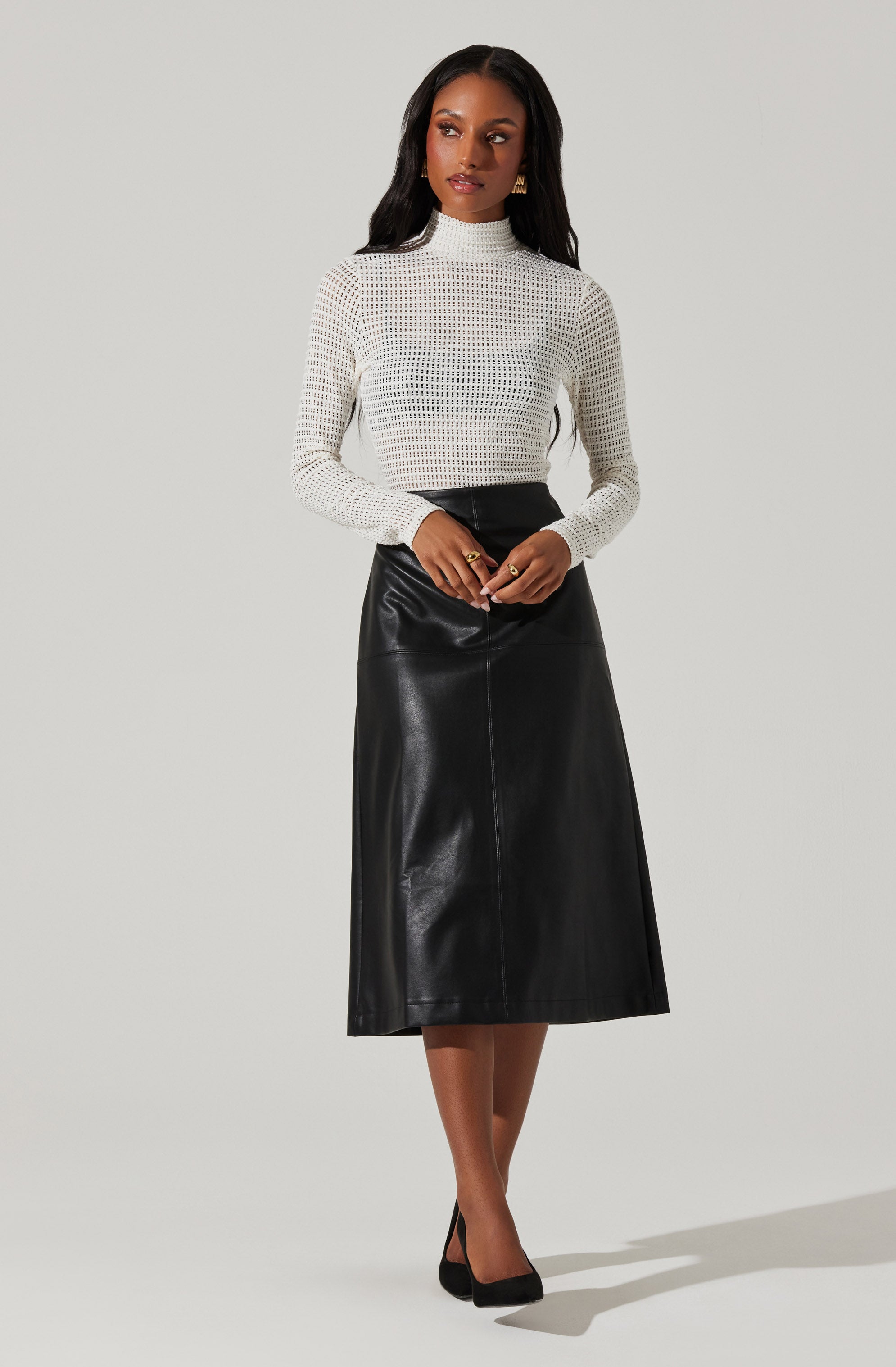A woman is wearing a white, long-sleeved, high-necked, open-knit sweater paired with a high-waisted, black, leather A-line midi skirt. She has black heels on and is accessorized with simple jewelry.