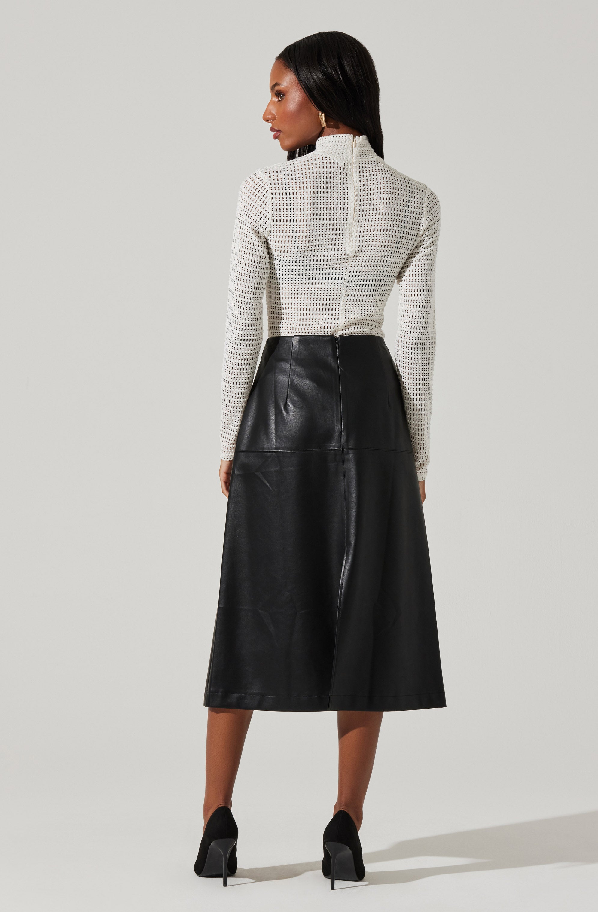 A woman is wearing a long-sleeved, white, open-knit top with a high neckline, paired with a high-waisted, black leather midi skirt that has a slight flare and a prominent back zipper. She is also wearing black high heels.