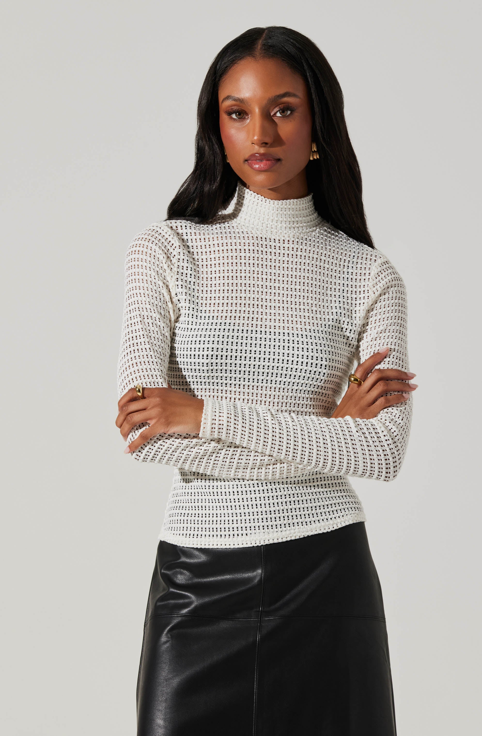 A woman is wearing a white, long-sleeved, high-neck knit sweater with a small, open weave pattern. She pairs it with a black, shiny, leather or faux leather skirt.
