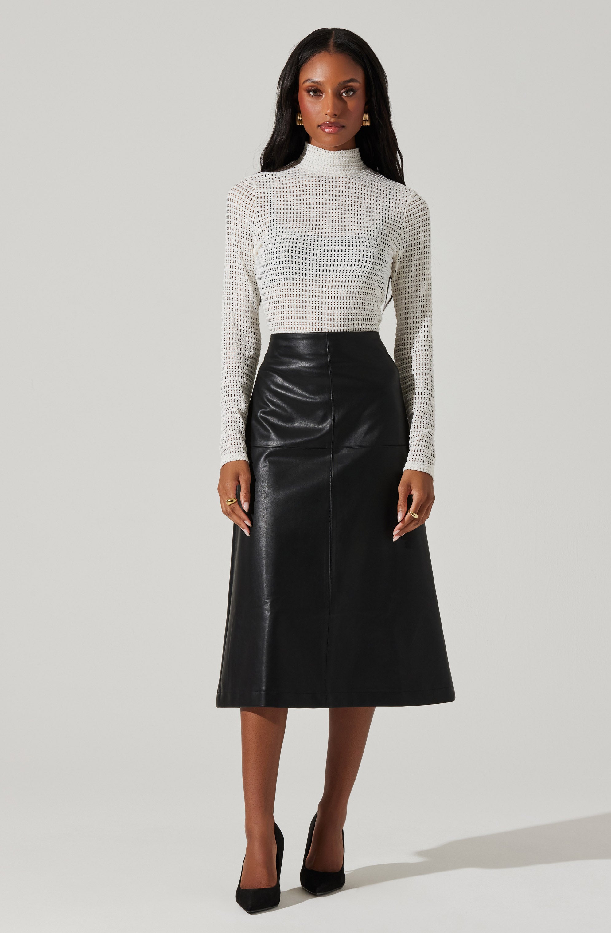 A woman is wearing a white, long-sleeved, high-neck top with a textured, open-knit pattern. She has paired it with a high-waisted, black leather midi skirt and black pointed-toe heels.