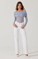 The woman is wearing an off-the-shoulder, long-sleeved top made of a textured, light blue fabric that is slightly see-through with a floral lace pattern. She is also wearing high-waisted, straight-leg white jeans with a button and zipper closure. She has on white heels with a pointed toe and a bow detail on the front.