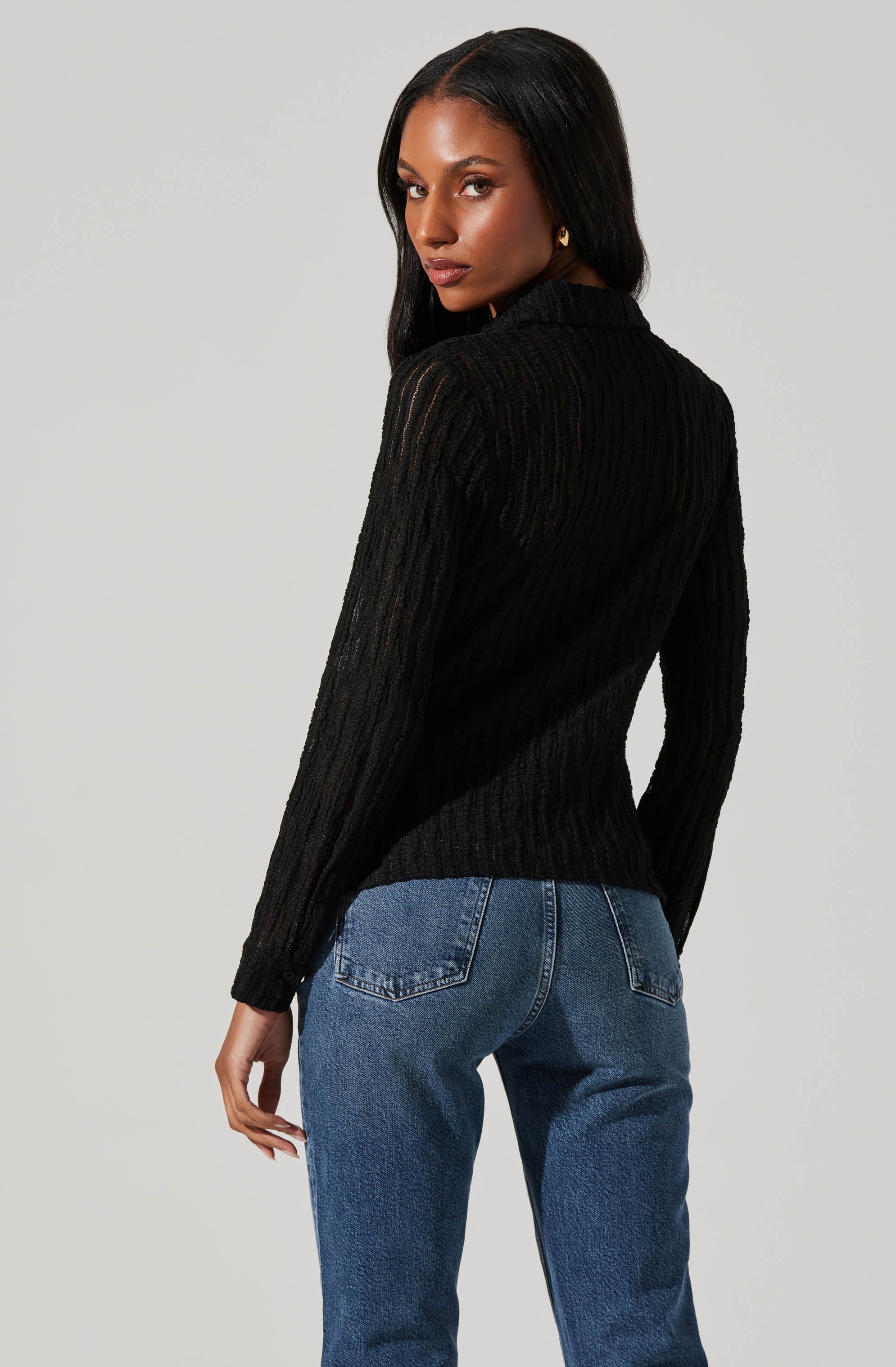 A woman is wearing a black, long-sleeved, ribbed knit sweater with a collared neckline, paired with blue jeans.