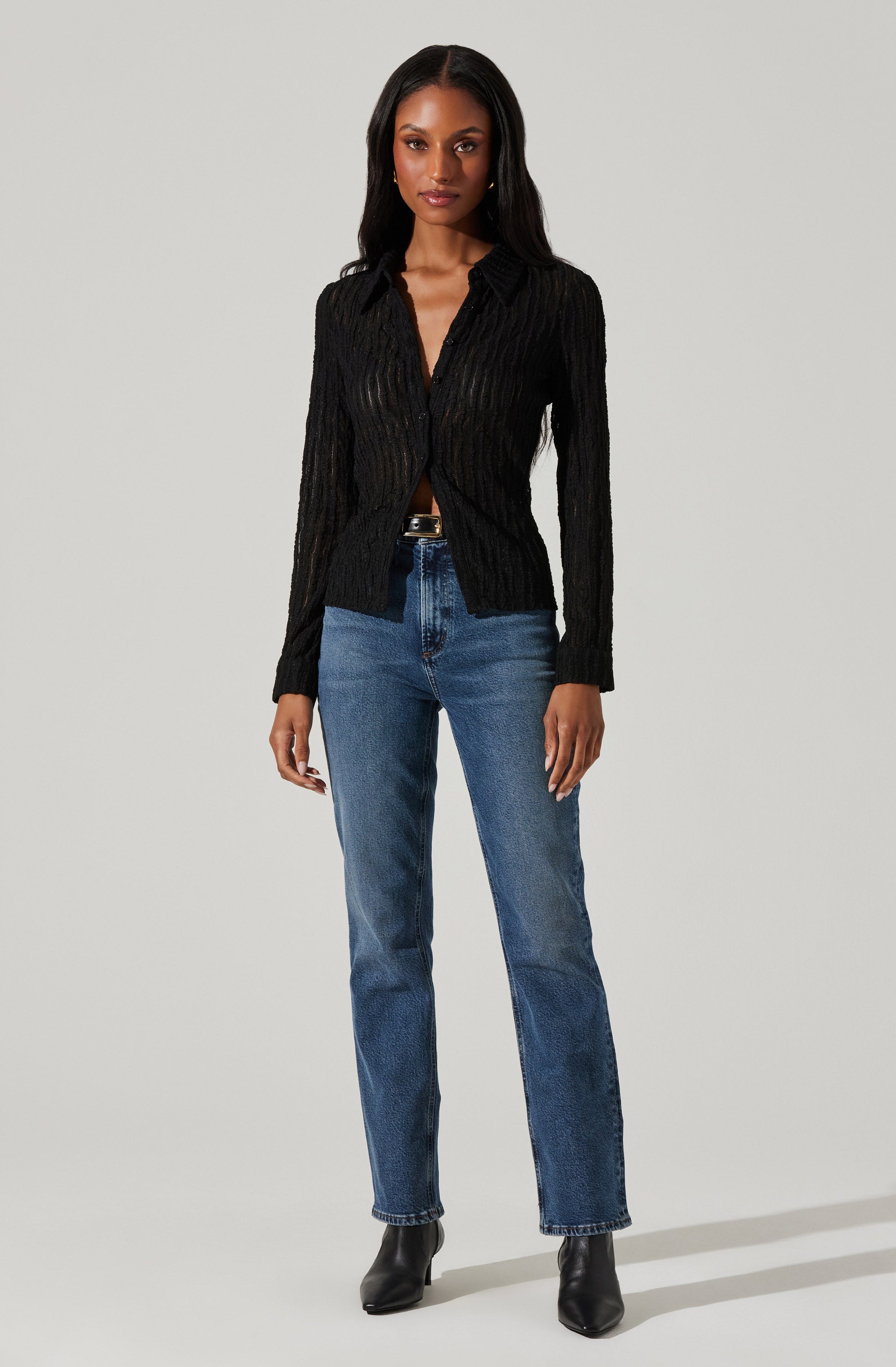 A woman is wearing a black, long-sleeved, textured sweater with a collar and a button-down front. She has paired it with blue high-waisted jeans and black ankle boots.