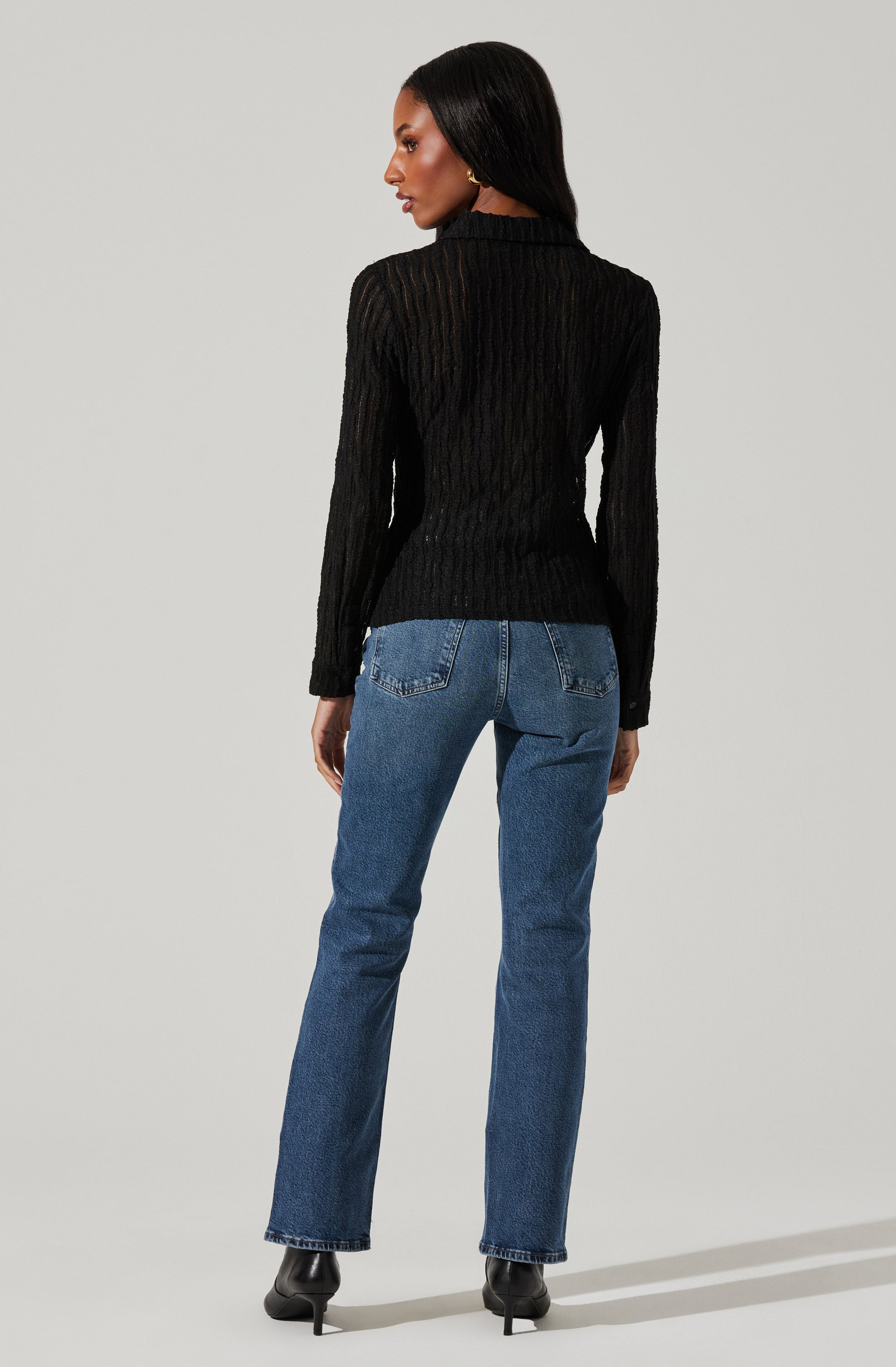 A woman is wearing a black, long-sleeved, textured sweater with a high neckline, paired with high-waisted blue jeans that have visible stitching and back pockets. She is also wearing black heeled ankle boots.
