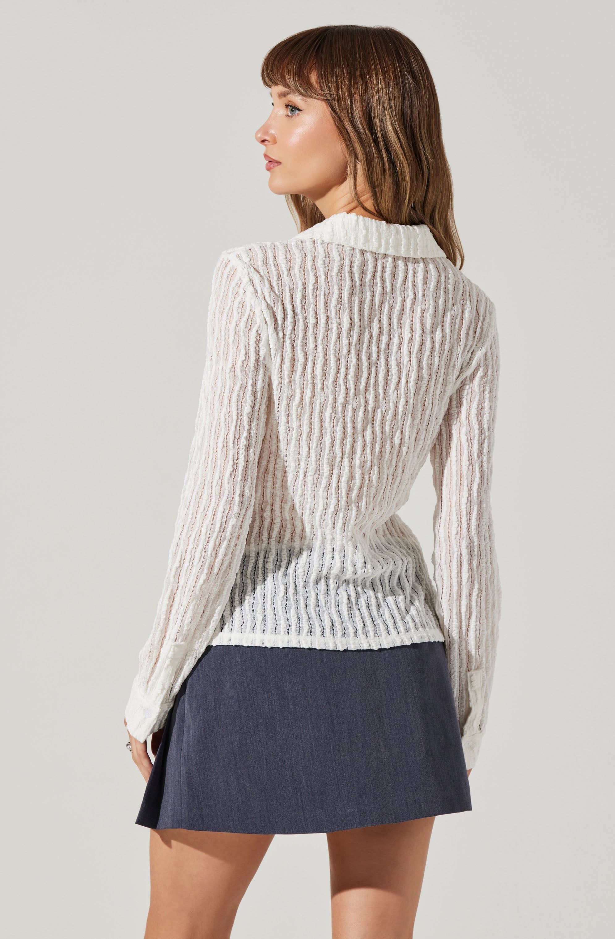 A woman is wearing a long-sleeved, cable-knit sweater with a turtleneck collar. The sweater is sheer with a textured pattern and is predominantly white with subtle beige or pinkish tones. She is also wearing a dark gray, A-line mini skirt made of a denim-like fabric.
