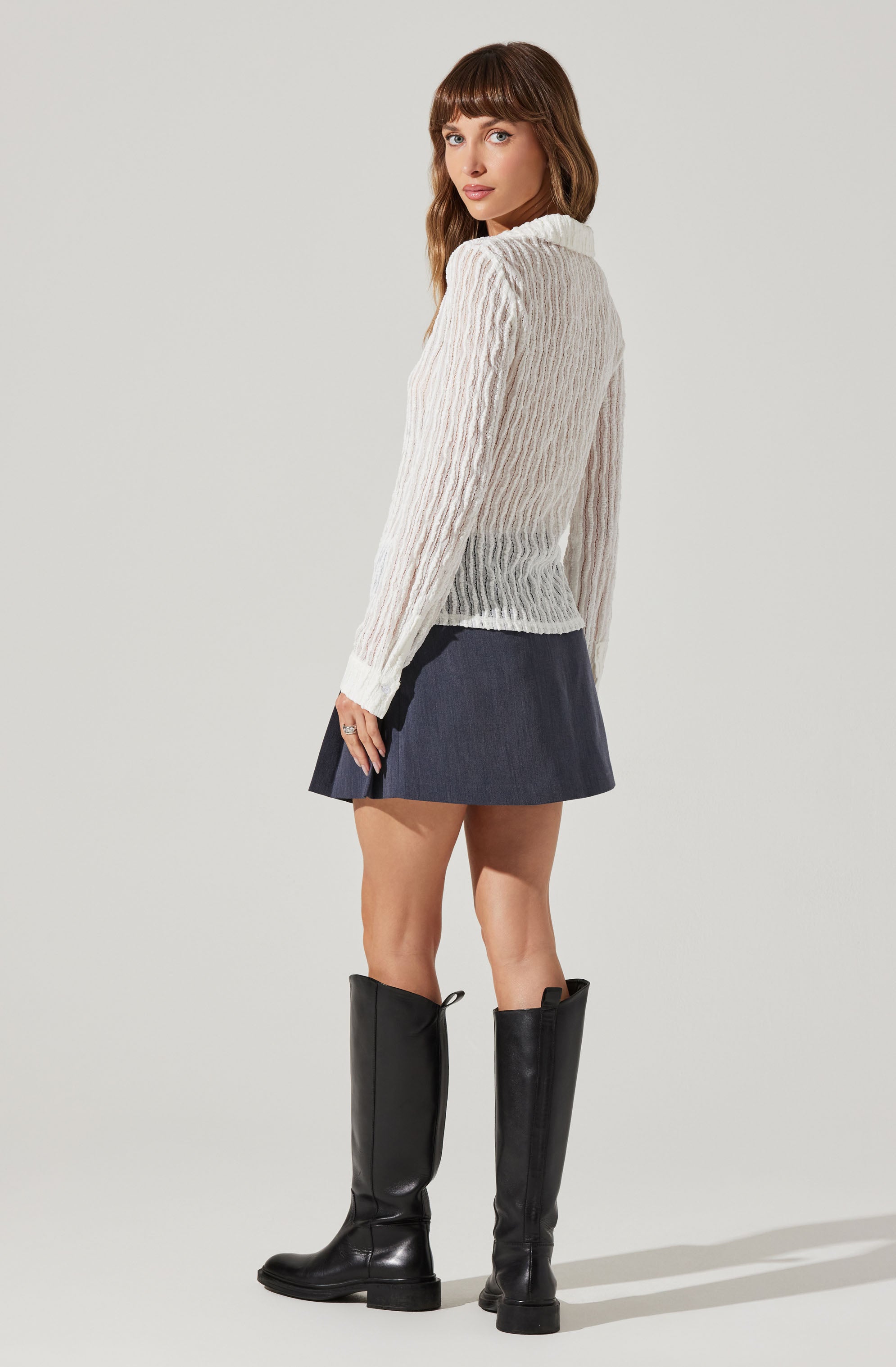 A woman is wearing a white, long-sleeved, cable-knit sweater with a high neckline. She has on a dark-colored, pleated, short skirt and tall black leather boots that reach just below the knee.