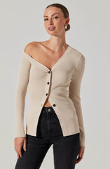A woman is wearing a beige, ribbed, V-neck button-up cardigan with long sleeves. The cardigan is slightly off-the-shoulder on her left side, and the buttons are dark-colored. She pairs the cardigan with high-waisted black jeans and has her hair styled in a sleek bun. She is also wearing large hoop earrings.