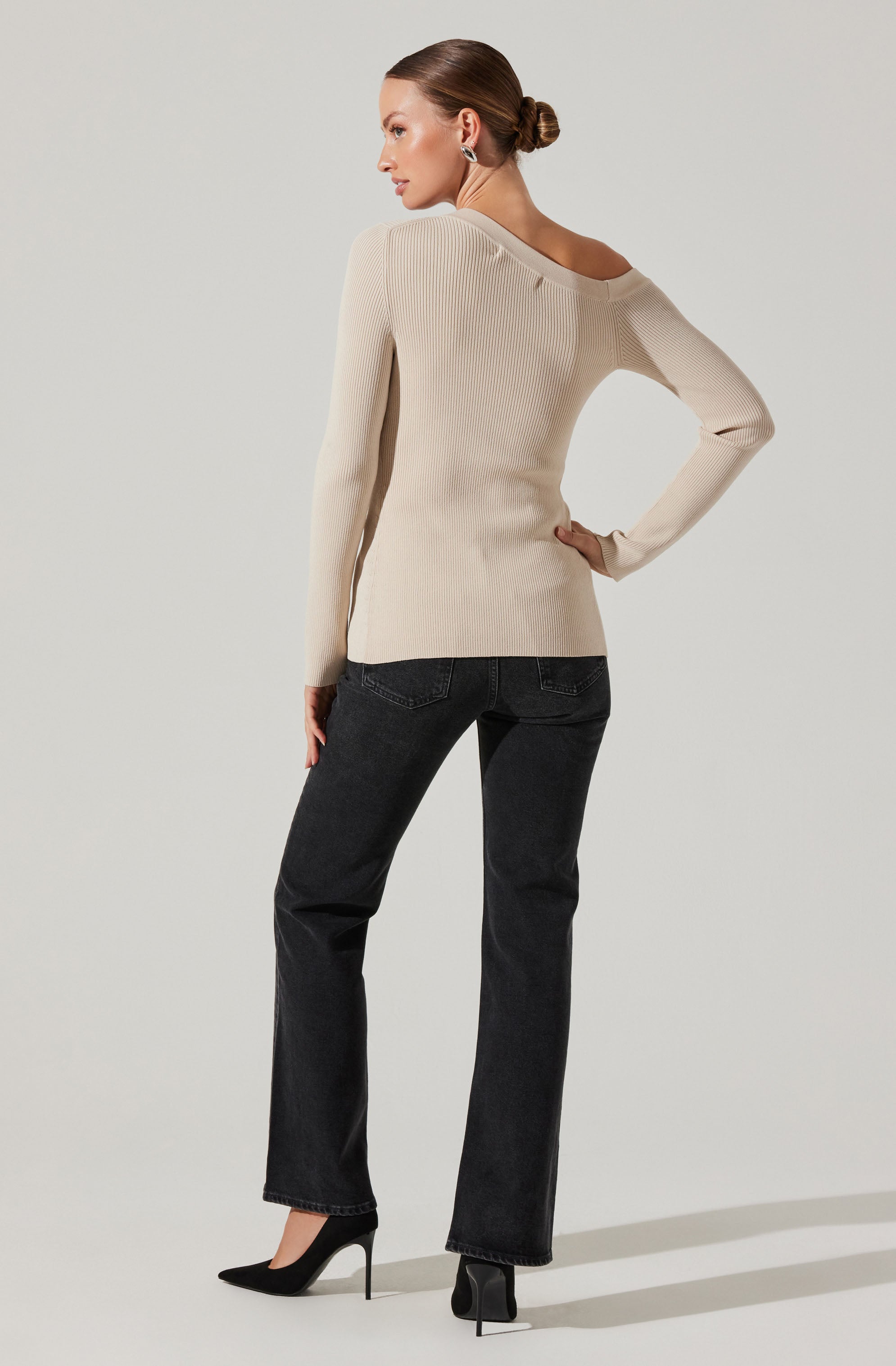 A woman is wearing a cream-colored, ribbed knit sweater with an asymmetrical neckline that falls off one shoulder. She pairs it with dark high-waisted jeans and black high-heeled shoes. Her hair is styled in a low bun, and she has on large earrings.