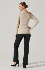 A woman is wearing a cream-colored, ribbed knit sweater with an asymmetrical neckline that falls off one shoulder. She pairs it with dark high-waisted jeans and black high-heeled shoes. Her hair is styled in a low bun, and she has on large earrings.