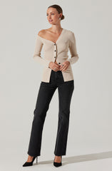 A woman is wearing a beige, long-sleeved, ribbed cardigan with black buttons down the front and an off-the-shoulder neckline. She pairs it with high-waisted, straight-leg black jeans and black pointed-toe high heels.