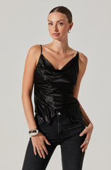 A woman is wearing a black satin camisole with thin spaghetti straps and a draped neckline. She pairs it with high-waisted black jeans. She accessorizes with a chunky silver bracelet on her left wrist, a silver ring on her right hand, and silver hoop earrings.