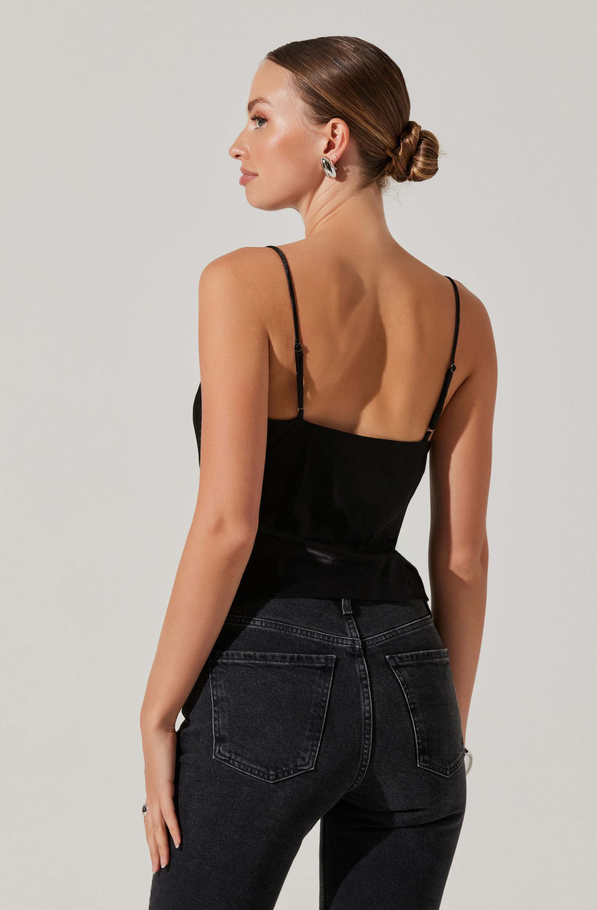 A woman is wearing a black spaghetti strap top with thin straps. She is also wearing high-waisted black jeans with visible stitching and back pockets. Her hair is styled in a low, twisted bun, and she has large hoop earrings.
