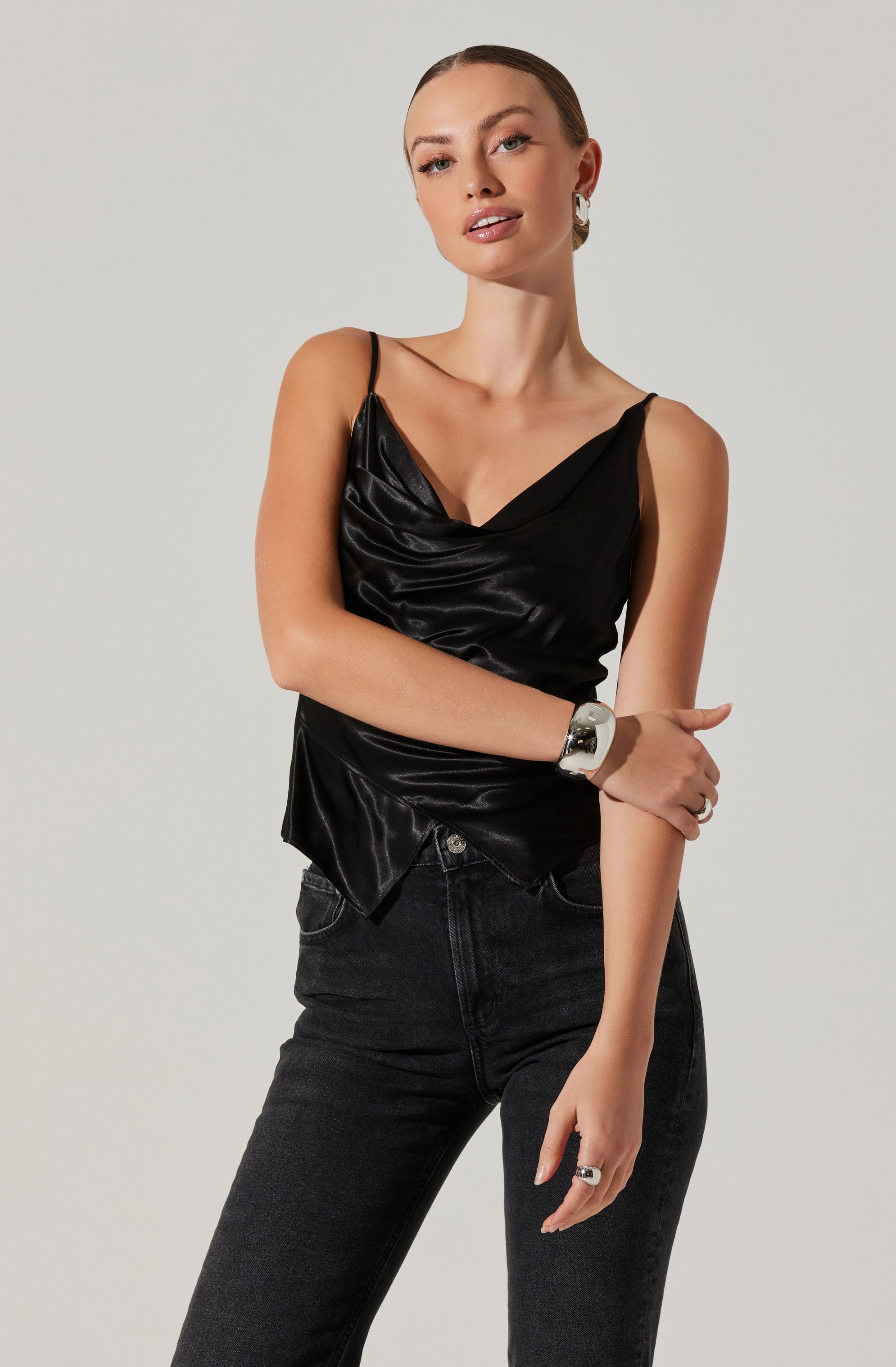 A woman is wearing a black satin camisole with thin spaghetti straps and a draped cowl neckline. She pairs it with high-waisted black jeans.